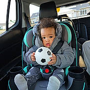 Enkore is registered to the contest to win money with this photo: baby, child, car_seat, toddler, soccer_ball, winter_clothing, jacket, seatbelt, car_interior, window, suburban, house, curly_hair, footwear, cozy, safety, curious, indoors, person, transportation