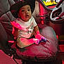 toddler, child, cowboy_hat, seat, chair, indoor, play_area, colorful_background, polka_dots, curious_expression, cushion, clothing, socks, hand_motion, steering_wheel, vehicle_interior, red_lighting, young_child, portrait, casual