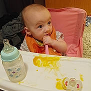 Miranda joined the competition — help win amazing prizes! baby, high_chair, spoon, food, messy, bib, bottle, pacifier, indoor, child, feeding, curious, pink, orange, table, utensil, infant, face, person, sitting