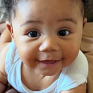 Lorenzo is registered to the contest to win money with this photo: baby, child, curly_hair, smiling, face, eyes, arm, skin, white_top, lying_down, soft_surface, portrait, cute, infant, closeup, person, happy, indoors, young_child, expression