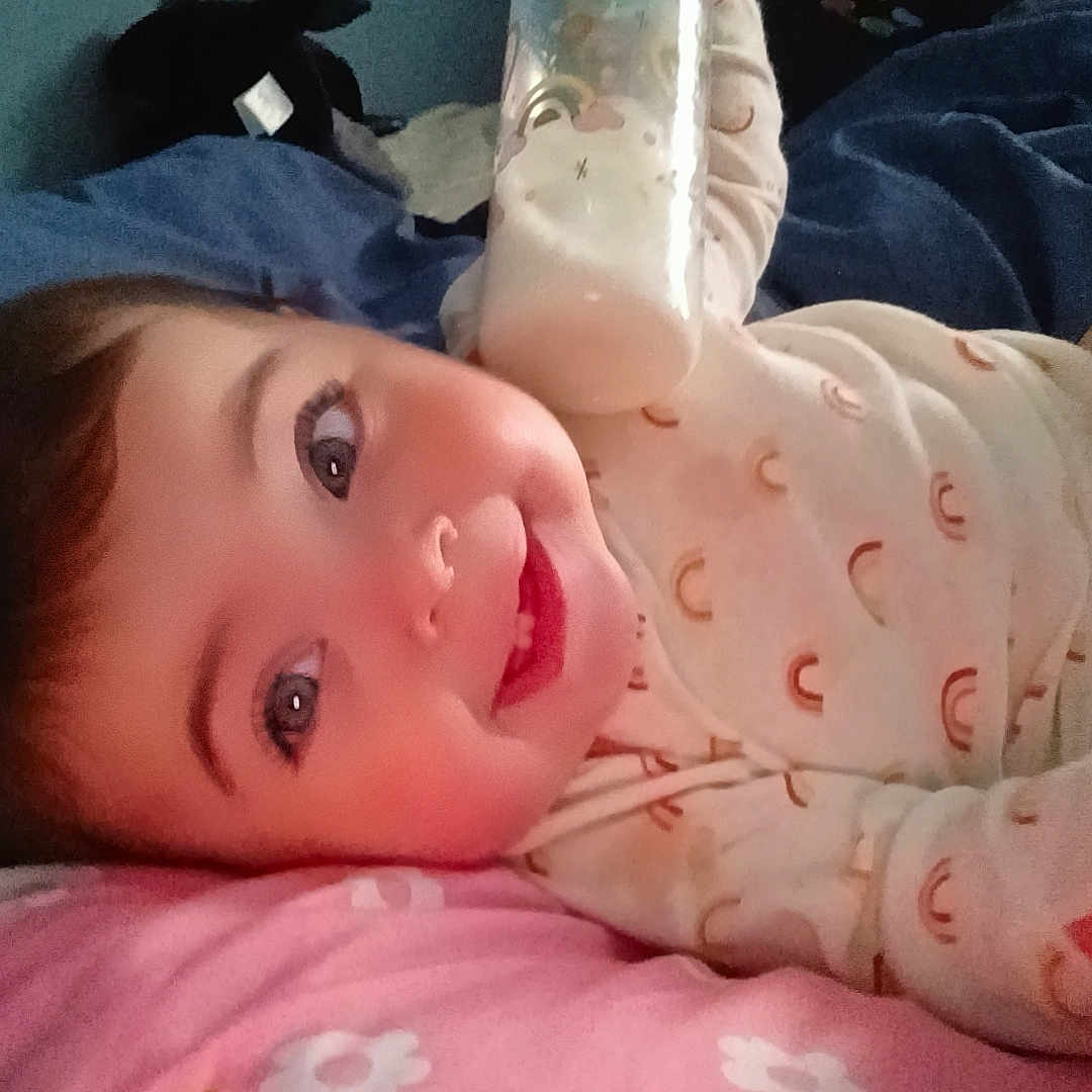 Adalynn joined the competition — help win amazing prizes! baby, smiling, bottle, milk, pillow, onesie, rainbow_pattern, bed, blanket, child, face, happy, indoor, person, infant, cute, lying_down, wall, blue_background, pink_pillow