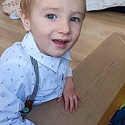 Logan a rejoint le concours — aidez-le/la à gagner de superbes lots ! toddler, child, indoor, cardboard_box, hair_spike, white_shirt, suspenders, floor, blanket, cushion, bed, hand, playing, expression, blue_eyes, light_skin, wooden_floor, casual_clothing, smiling, portrait