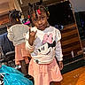child, toddler, girl, portrait, mirror, reflection, braids, hair_beads, smiling, hand_gesture, minnie_mouse, shirt, skirt, socks, shoes, indoors, bedroom, wood_floor, furniture, colorful_clothing