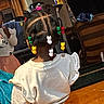 child, toddler, back_view, braids, beads, colorful_hair_accessories, white_shirt, pink_pants, sitting, indoors, living_room, television, laundry, furniture, wood_floor, chair, household, portrait, casual_clothing, family_home