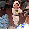 child, toddler, smiling, happy, hoodie, casual_clothing, standing, restaurant, dining_area, chairs, tables, tablecloth, indoor, portrait, playful, funny_pose, dark_skin, curly_hair, patterned_fabric, photograph