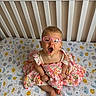 Serenity is registered to the contest to win money with this photo: baby, child, glasses, pink_glasses, floral_dress, crib, crib_slats, patterned_sheet, sun_pattern, moon_pattern, star_pattern, sitting, expression, mouth_open, indoor, nursery, colorful, cute, person, young_child