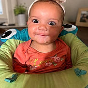 Kaizlynn joined the competition — help win amazing prizes! baby, child, infant, smiling, headband, orange_shirt, flower_print, cushioned_seat, indoor, portrait, wide_eyes, cute, cheeky, happy, person, face, head, baby_seat, colorful, home