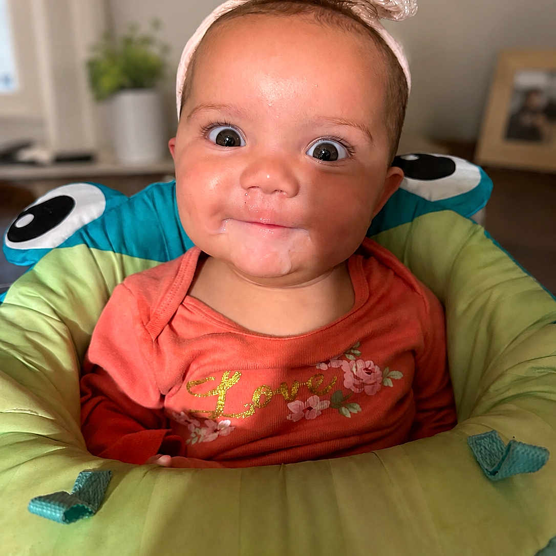Kaizlynn joined the competition — help win amazing prizes! baby, child, infant, smiling, headband, orange_shirt, flower_print, cushioned_seat, indoor, portrait, wide_eyes, cute, cheeky, happy, person, face, head, baby_seat, colorful, home
