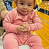 baby, child, pink_outfit, yellow_bow, headband, smile, hands, gymnasium, wooden_floor, people, background, seated, cushion, indoor, casual_clothing, curious_expression, infant, portrait, colorful, young_child