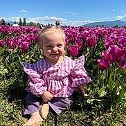 Gwendolyn joined the competition — help win amazing prizes! toddler, child, flower_field, tulips, pink_flowers, outdoor, nature, sunny_day, grass, greenery, smiling, cute, pigtails, purple_pants, striped_shirt, baby_shoes, plants, blue_sky, mountains, happy