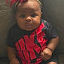baby, infant, headband, red_bow, nike, onesie, black_clothing, red_clothing, couch, seated, chubby_cheeks, curious_expression, indoor, person, skin, hand, legs, furniture, portrait, closeup