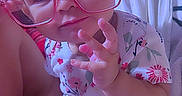 Raven is registered to the contest to win money with this photo: baby, bed, blanket, casual_photo, child, close_up, colorful_lighting, face, floral_clothing, glasses, hand_gesture, heart_sticker, indoors, infant, onesie, pink_glasses, portrait, sitting, smiling, timestamp