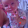 baby, bed, blanket, casual_photo, child, close_up, colorful_lighting, face, floral_clothing, glasses, hand_gesture, heart_sticker, indoors, infant, onesie, pink_glasses, portrait, sitting, smiling, timestamp