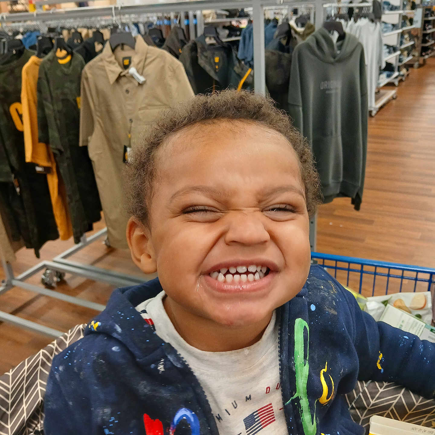 Kyrie Amir is registered to the contest to win money with this photo: boy, casual_clothing, child, clothing, curly_hair, face, grocery, happy, hoodie, indoor, person, rack, retail, shelf, shopping_cart, smiling, store, teeth, tshirt, wood_floor