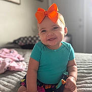 Jaylani is registered to the contest to win money with this photo: baby, bed, blanket, child, cute, furniture, hand_holding, happy, headband, home, indoor, infant, orange_bow, person, portrait, skin, skirt, smile, soft_light, teal_shirt