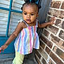 toddler, child, girl, curly_hair, colorful_clothing, striped_top, yellow_pants, outdoor, brick_wall, blue_door, side_view, walking, reaching, cute, young_child, person, portrait, daylight, casual, standing