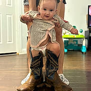 Cora joined the competition — help win amazing prizes! baby, child, cowboy_boots, diaper, indoor, person, wooden_floor, holding_hands, smiling, cute, home, adult, legs, footwear, toy, furniture, living_room, shorts, playful, funny