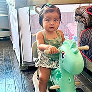 Adeline is registered to the contest to win money with this photo: toddler, child, toy, unicorn, ride_on, indoor, floor, playpen, curtain, window, couch, quilt, hair_tie, sandal, green_top, skirt, wooden_floor, face, person, furniture