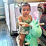 toddler, child, toy, unicorn, ride_on, indoor, floor, playpen, curtain, window, couch, quilt, hair_tie, sandal, green_top, skirt, wooden_floor, face, person, furniture