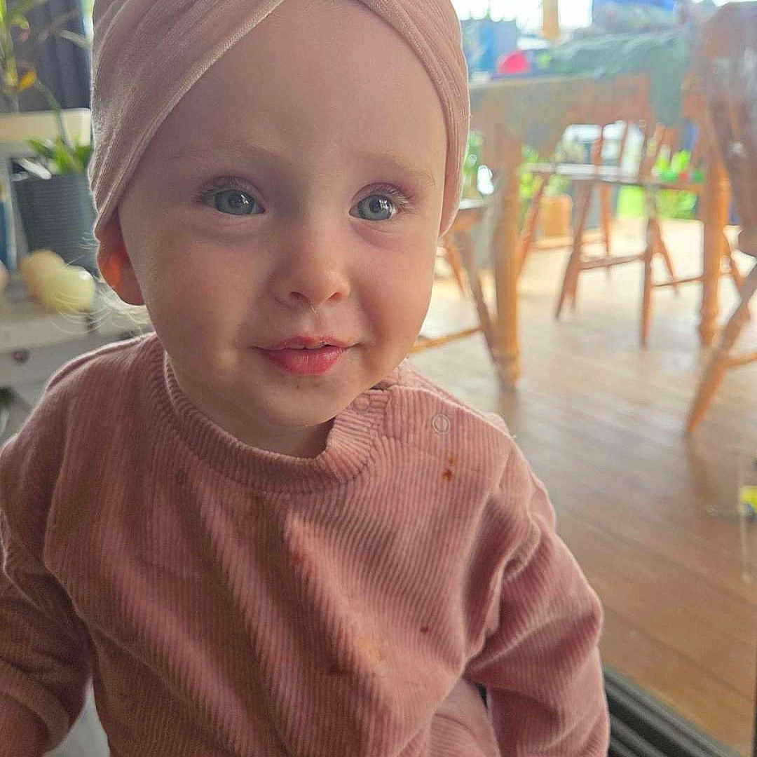 Allora joined the competition — help win amazing prizes! bow, casual_wear, child, closeup, curious_expression, face, floor, headband, home, indoor, long_sleeves, natural_light, pants, person, pink_clothing, plant, portrait, toddler, wooden_furniture, young_child