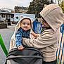 baby, blanket, casual, child, clothing, community_center, fence, happy, holding, hoodie, interaction, outdoor, park, person, playground, smile, stroller, toddler, trees, young