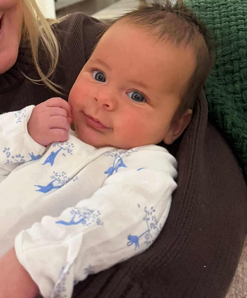 Callum joined the competition — help win amazing prizes! baby, infant, child, face, eyes, smiling, onesie, hand, adult_partial, cuddle, blanket, knit_blanket, indoor, portrait, cozy, couch, blue_pattern, skin, hair, adorable