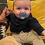 baby, pacifier, child, couch, hand, pillow, clothing, indoor, person, infant, sitting, cute, young_child, patterned_pants, smiling_eyes, holding_hand, yellow_pillow, dark_shirt, home, cozy