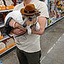 man, dog, cowboy_hat, pet, store, aisle, shelf, products, casual_clothing, beard, leash, holding, small_dog, wooden_crafts, shopping_cart, floor, indoor, retail, tags, curious