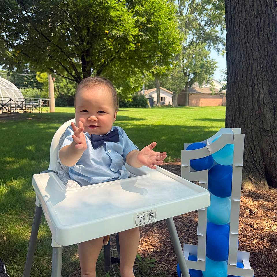 Alim is registered to the contest to win money with this photo: baby, high_chair, blue_bow_tie, smiling, outdoor, grass, tree, balloons, number_one, birthday, child, sunlight, celebration, greenery, footwear, happy, person, clothing, daytime, park