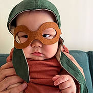 Alim is registered to the contest to win money with this photo: baby, child, face, hand, goggles, hat, green_hat, orange_shirt, person, infant, indoors, couch, holding, cute, portrait, expression, costume, headwear, closeup, skin