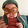baby, child, face, hand, goggles, hat, green_hat, orange_shirt, person, infant, indoors, couch, holding, cute, portrait, expression, costume, headwear, closeup, skin