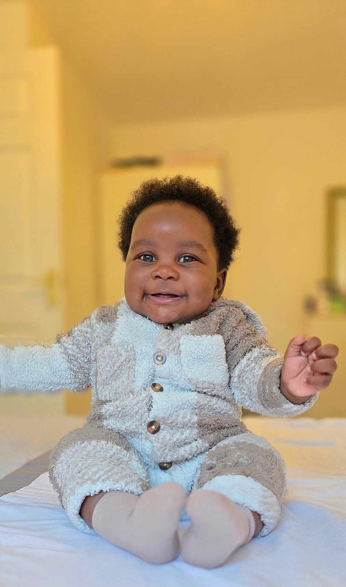 Rukudzo Wesley is registered to the contest to win money with this photo: baby, child, smiling, sitting, indoors, cozy_clothing, fuzzy_outfit, bed, happy, cute, person, infant, portrait, warm_lighting, home, soft_texture, hands, feet, face, curly_hair