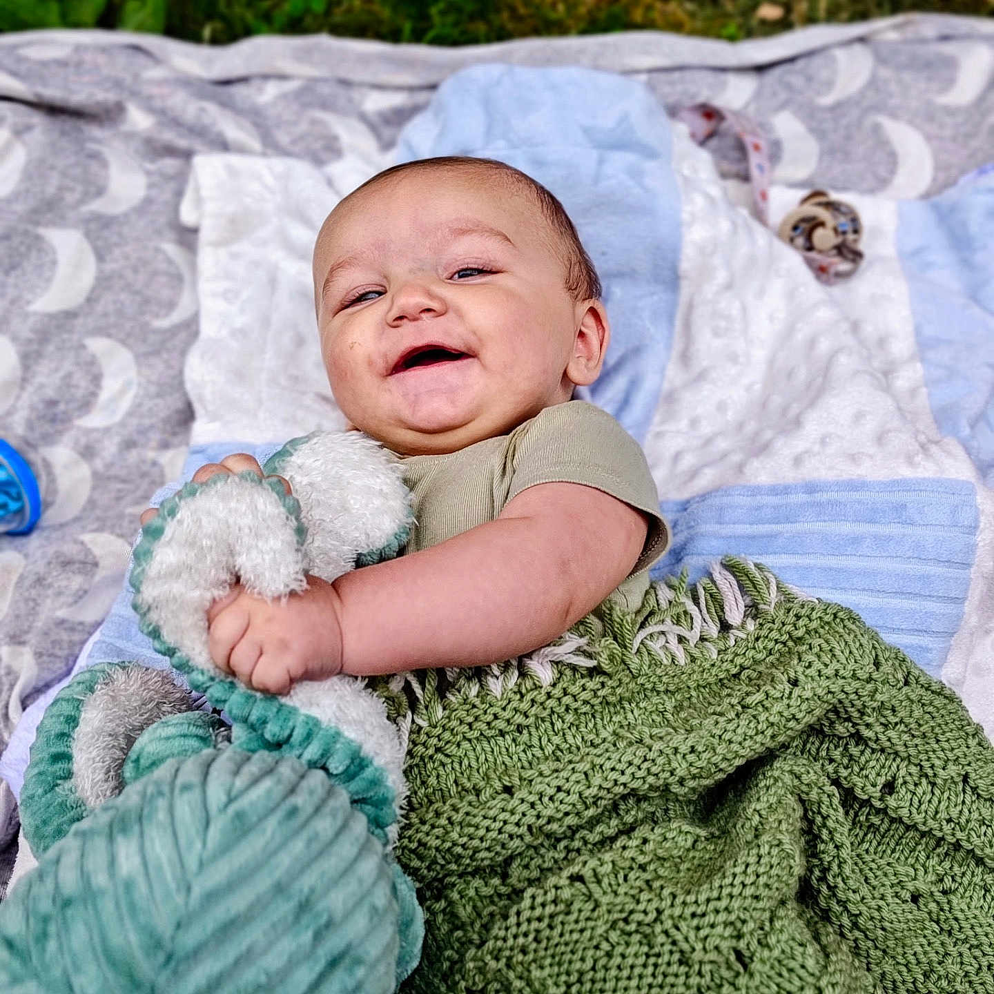 Westley is registered to the contest to win money with this photo: baby, infant, blanket, plush_toy, smiling, outdoor, grass, quilt, knit, cute, happy, child, toy, soft_texture, lying_down, green, blue, white, face, person