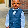 child, boy, smile, formal_wear, vest, bow_tie, white_shirt, portrait, standing, outdoor, confident, short_hair, fashion, cute, person, young, happy, background_blur, brick_wall, hands