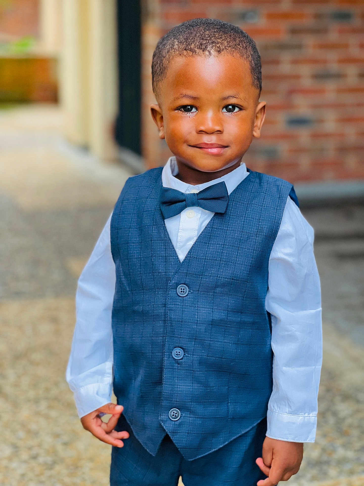 Sekou participe au concours pour gagner de l'argent avec cette photo : child, boy, smile, formal_wear, vest, bow_tie, white_shirt, portrait, standing, outdoor, confident, short_hair, fashion, cute, person, young, happy, background_blur, brick_wall, hands