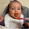 baby, child, high_chair, bib, baby_food, spoon, messy_face, tongue, purple_bows, feeding_time, infant, cute, face, clothing, person, indoor, expression, young_child, table, feeding