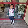 child, girl, smiling, backpack, white_vest, green_sweater, blue_skirt, white_tights, black_shoes, braided_hair, bracelet, potted_plant, glass_door, reflection, floor_tiles, indoors, entrance, happy, person, portrait