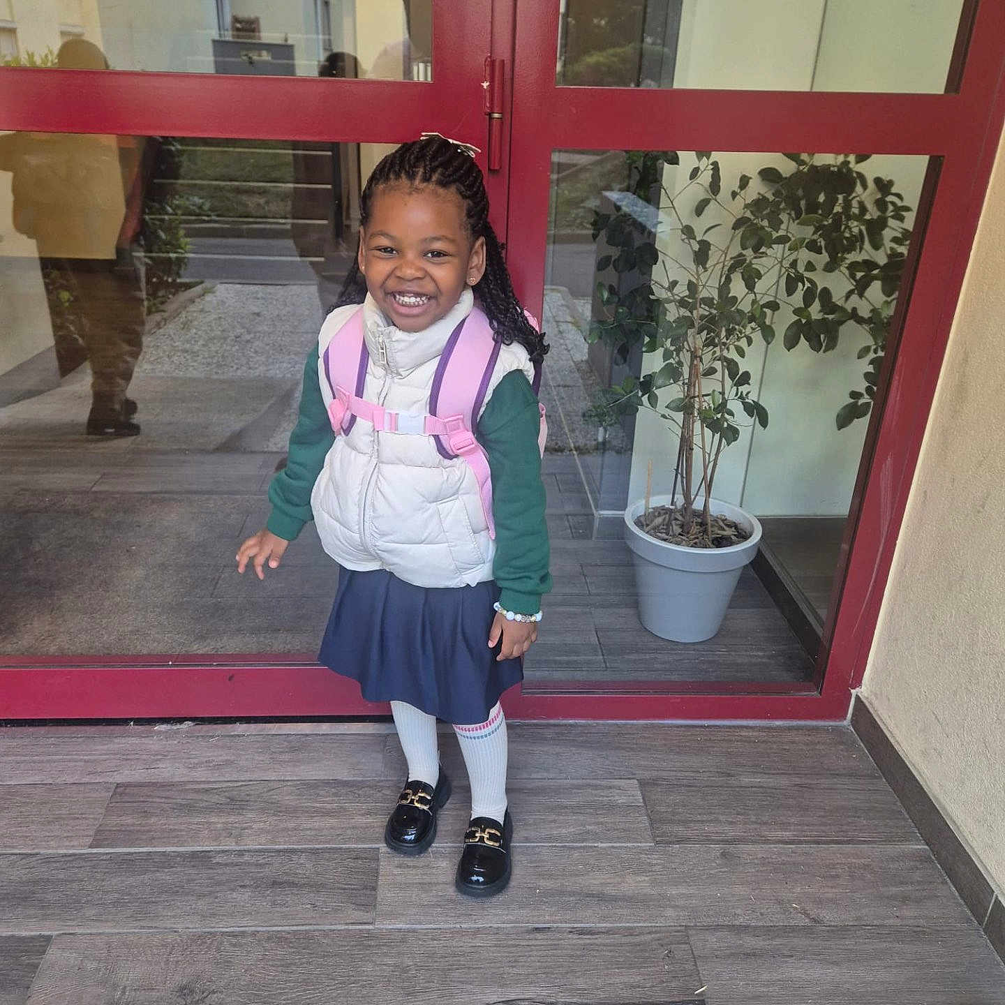 Imya participe au concours pour gagner de l'argent avec cette photo : backpack, black_shoes, blue_skirt, bracelet, braided_hair, child, entrance, floor_tiles, girl, glass_door, green_sweater, happy, indoors, person, portrait, potted_plant, reflection, smiling, white_tights, white_vest