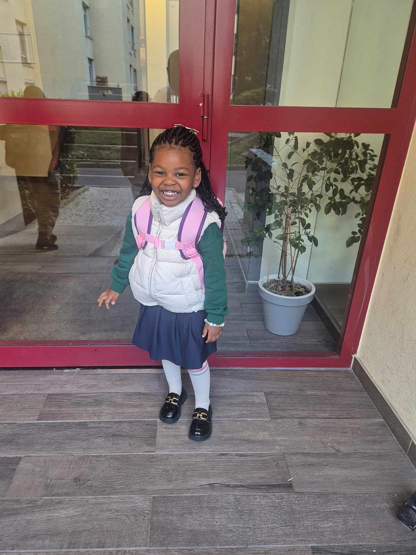 Imya participe au concours pour gagner de l'argent avec cette photo : child, girl, smiling, backpack, white_vest, green_sweater, blue_skirt, white_tights, black_shoes, braided_hair, bracelet, potted_plant, glass_door, reflection, floor_tiles, indoors, entrance, happy, person, portrait