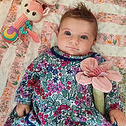 Elina participe au concours pour gagner de l'argent avec cette photo : baby, child, blue_eyes, floral_dress, plush_toy, pink_flower, blanket, patterned_blanket, soft_toy, indoor, cute, smiling, laying_down, infant, person, portrait, colorful, young_child, cozy, playtime