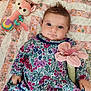 baby, child, blue_eyes, floral_dress, plush_toy, pink_flower, blanket, patterned_blanket, soft_toy, indoor, cute, smiling, laying_down, infant, person, portrait, colorful, young_child, cozy, playtime