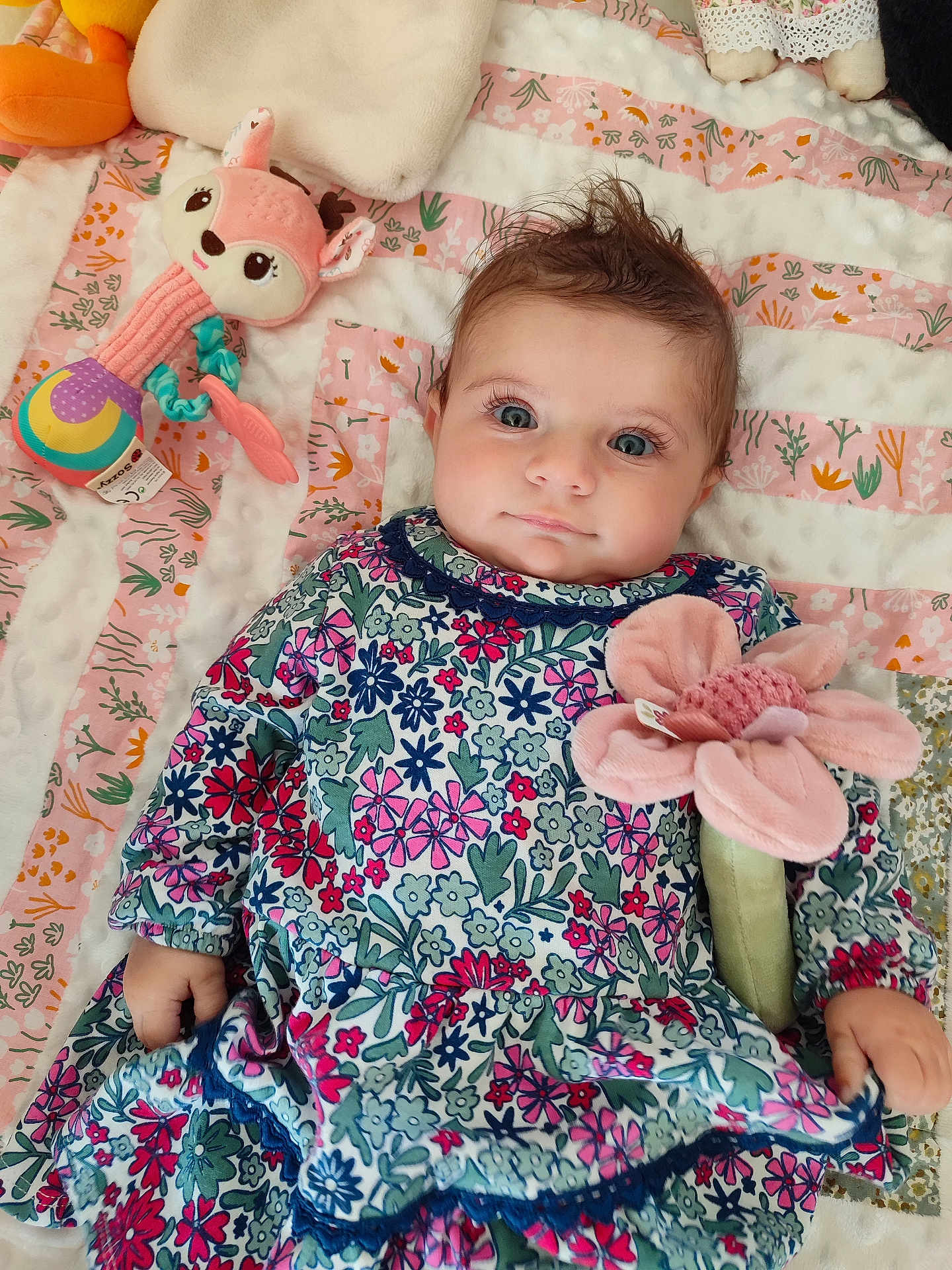 Elina participe au concours pour gagner de l'argent avec cette photo : baby, child, blue_eyes, floral_dress, plush_toy, pink_flower, blanket, patterned_blanket, soft_toy, indoor, cute, smiling, laying_down, infant, person, portrait, colorful, young_child, cozy, playtime