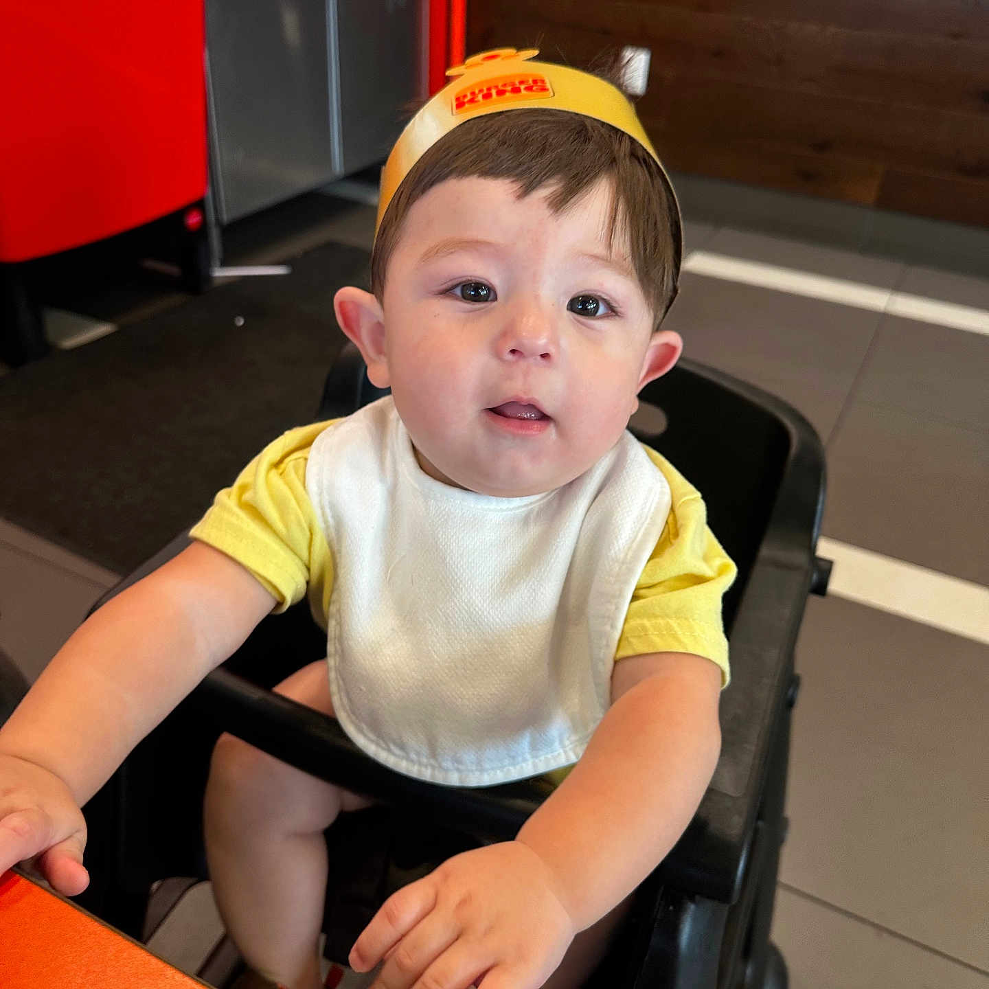 Rogelio joined the competition — help win amazing prizes! baby, bib, brown_hair, child, curious, expression, face, floor, furniture, hand, high_chair, indoor, person, restaurant, seated, short_sleeves, skin, table, yellow_crown, young