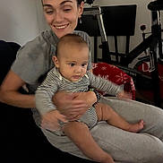 Nathaniel is registered to the contest to win money with this photo: woman, baby, indoor, striped_clothing, smile, exercise_bike, blanket, holding, seated, person, happy, casual_clothing, home, adult, child, family, comfort, toddler, living_room, relaxed