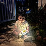Eliot is registered to the contest to win money with this photo: child, curious, evening, fence, glow, grass, lantern, light, night, outdoor, person, plants, portrait, shadow, shirt, shoes, shorts, sidewalk, sitting, toddler