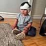 baby, casual_clothing, child, cozy, curious, floor, footwear, grey_shirt, headgear, helmet, home, indoor, person, rug, shoes, shorts, sitting, toddler, wooden_floor, young_child