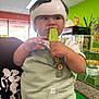 bracelet, bright_colors, button, child, clothing, counter, face, food, green_popsicle, hand, helmet, indoor, person, plant, popsicle, shorts, snack, store, toddler, window