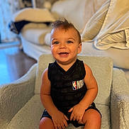 Michael is registered to the contest to win money with this photo: toddler, child, smiling, sitting, nba_jersey, black_clothing, moawk_hair, indoor, chair, couch, fan, happy, cute, person, portrait, home, furniture, wooden_floor, baby, small_hands