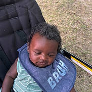 Steffon Cox joined the competition — help win amazing prizes! baby, infant, child, stroller, bib, sleepy, portrait, outdoor, grass, seated, harness, straps, curly_hair, facial_expression, clothing, green_outfit, blue_bib, photography, closeup, park