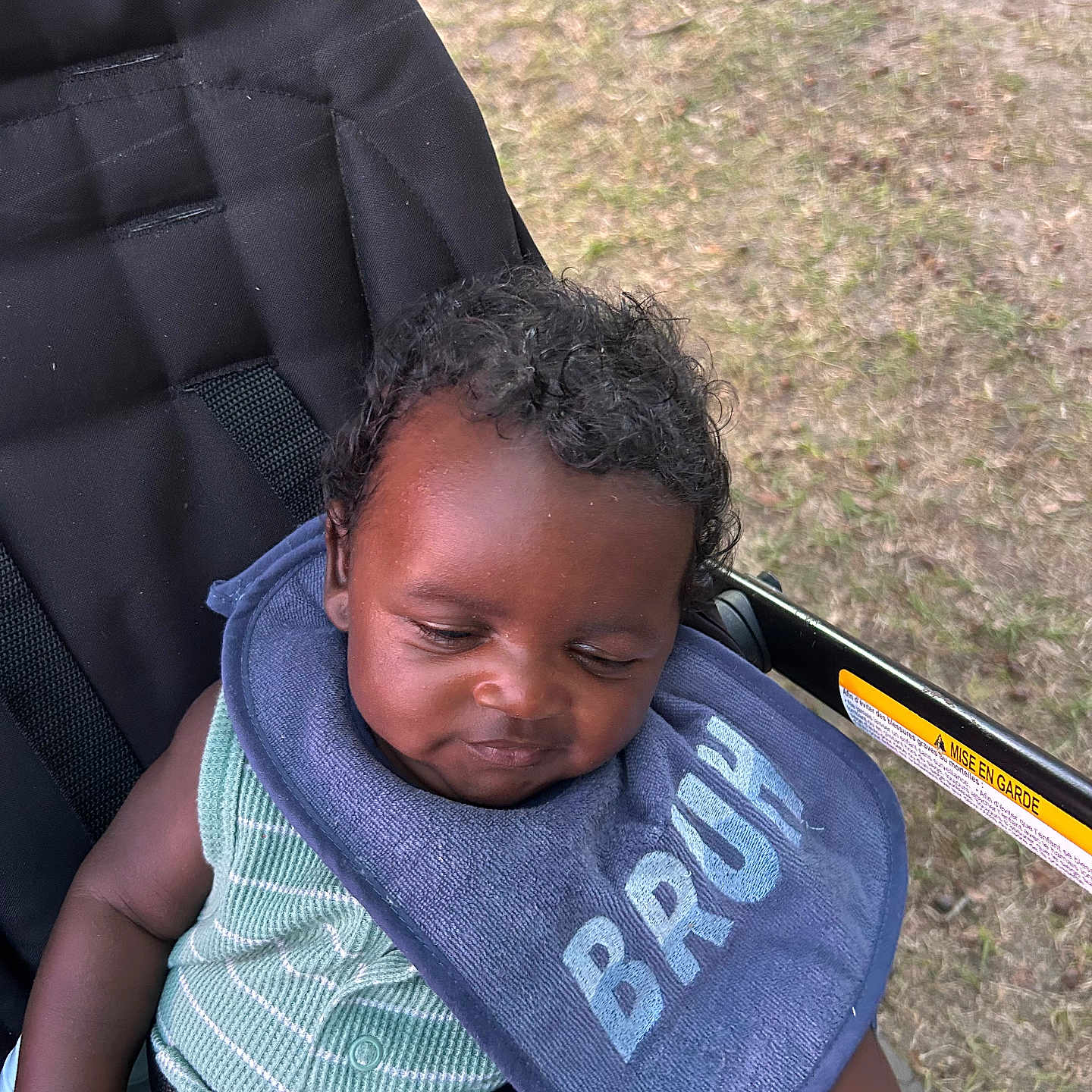 Steffon Cox joined the competition — help win amazing prizes! baby, infant, child, stroller, bib, sleepy, portrait, outdoor, grass, seated, harness, straps, curly_hair, facial_expression, clothing, green_outfit, blue_bib, photography, closeup, park